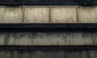 A close-up view of an industrial metal structure featuring rusted steel beams with rivets and a weathered, textured surface. The beams have a greenish, corroded appearance, suggesting wear and age.