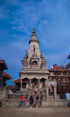 A stone temple structure with intricate carvings, flanked by tourists who are exploring and taking pictures. The temple is surrounded by some traditional buildings, and there is a clear blue sky in the background. Scaffolding can be seen on the right side, indicating some construction or restoration work.