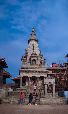 A stone temple structure with intricate carvings, flanked by tourists who are exploring and taking pictures. The temple is surrounded by some traditional buildings, and there is a clear blue sky in the background. Scaffolding can be seen on the right side, indicating some construction or restoration work.