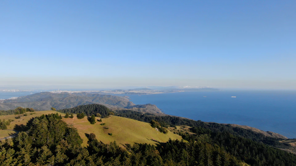 The image presents a breathtaking aerial view of a verdant hillside, with a body of water nestled at its base and a distant island visible on the horizon. The hill is blanketed in lush greenery, dotted with trees that add depth to the scene. The sky above mirrors the color of the sea below, creating a harmonious blend of natural elements. The perspective from which the photo was taken gives an expansive view of this picturesque landscape, inviting viewers to imagine themselves standing on top of the hill and taking in the panoramic vista.