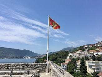 A red flag with a coat of arms flutters on a flagpole against a backdrop of a scenic coastal town. The town features buildings with red-tiled roofs nestled among green hills. A stone wall and railing are visible in the foreground, overlooking a body of water with mountains in the distance.