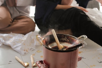 A close-up view of a traditional wax pot used for batik making is placed on the floor. It is surrounded by spilt wax and cloths stained with wax patterns. People sit around the pot, partially visible with their hands and laps suggesting they are engaged in batik crafting.