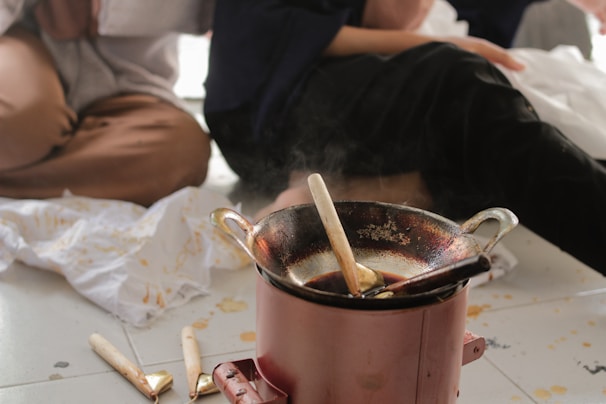 A close-up view of a traditional wax pot used for batik making is placed on the floor. It is surrounded by spilt wax and cloths stained with wax patterns. People sit around the pot, partially visible with their hands and laps suggesting they are engaged in batik crafting.