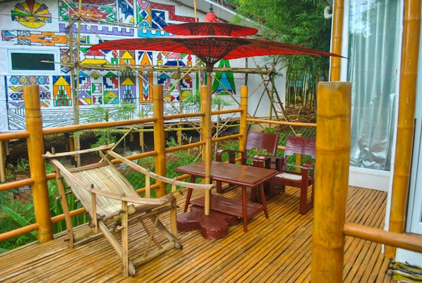A bamboo patio with wooden furniture is set against a background of a vibrant, geometric mural. There is a wooden hammock, a red umbrella providing shade, and a small wooden table with chairs. The area is surrounded by greenery and bamboo railings, creating a serene and natural atmosphere.