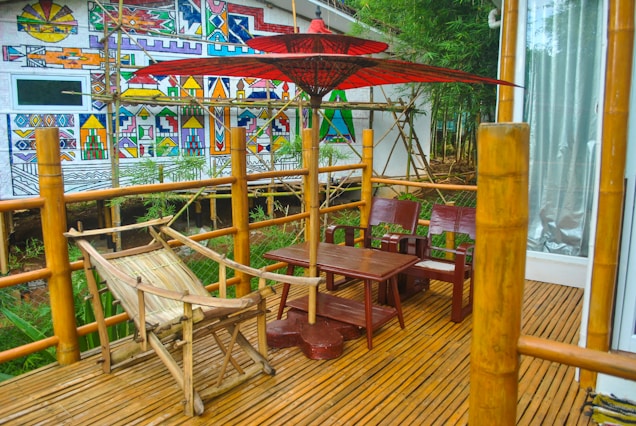 A bamboo patio with wooden furniture is set against a background of a vibrant, geometric mural. There is a wooden hammock, a red umbrella providing shade, and a small wooden table with chairs. The area is surrounded by greenery and bamboo railings, creating a serene and natural atmosphere.