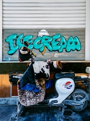 A motorbike covered with blankets and a jacket is parked against a shutter with vibrant graffiti spelling 'Ice Cream' in large teal letters with a stylized ice cream cone. The shutter is partially open and the scene suggests a casual and urban setting.