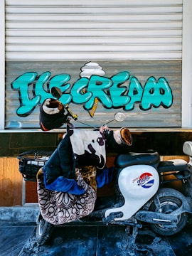 A motorbike covered with blankets and a jacket is parked against a shutter with vibrant graffiti spelling 'Ice Cream' in large teal letters with a stylized ice cream cone. The shutter is partially open and the scene suggests a casual and urban setting.
