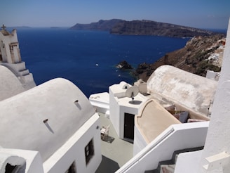 A coastal scene featuring traditional whitewashed buildings with smooth, curved rooftops and a deep blue sea in the background. The landscape includes a rocky shoreline and distant islands under a clear sky, conveying a serene Mediterranean atmosphere.