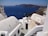 A coastal scene featuring traditional whitewashed buildings with smooth, curved rooftops and a deep blue sea in the background. The landscape includes a rocky shoreline and distant islands under a clear sky, conveying a serene Mediterranean atmosphere.