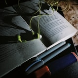 An open book is partially illuminated, with a pair of green earphones resting on its pages. Nearby, there are two pens and a highlighter pen, all placed on a dark surface. The lighting creates dramatic shadows, enhancing the contrast between light and dark areas.