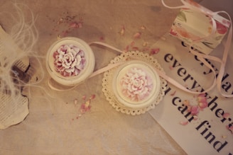 A soft, vintage-style arrangement featuring two candles in delicate glass holders with intricate flower designs on top. The candles sit on a beige surface, accompanied by scattered pink rose petals. To the side, there is a piece of paper with partly visible text, a lace doily, and a decorative gift box with floral patterns and a pink ribbon.