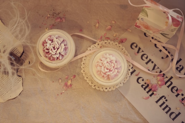 A soft, vintage-style arrangement featuring two candles in delicate glass holders with intricate flower designs on top. The candles sit on a beige surface, accompanied by scattered pink rose petals. To the side, there is a piece of paper with partly visible text, a lace doily, and a decorative gift box with floral patterns and a pink ribbon.