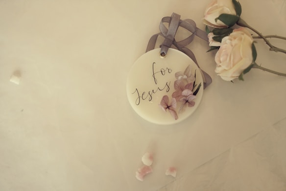 A decorative tag with the words 'for Jesus' written on it, adorned with pastel-colored flowers and a ribbon, lies on a light surface next to a bunch of soft pink roses.
