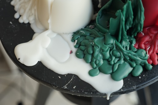 Melted wax in various colors, primarily white, green, and red, pooled and mixed on a dark surface. The wax appears soft and malleable, forming organic, flowing shapes.