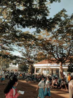 A group of children in school uniforms, many wearing masks, walk and gather in a schoolyard shaded by large trees. Numerous motorcycles are parked near a school building, with more people visible in the background. The atmosphere is lively and busy, with a basketball hoop visible in the distance.