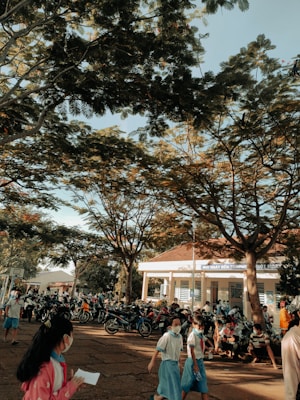 A group of children in school uniforms, many wearing masks, walk and gather in a schoolyard shaded by large trees. Numerous motorcycles are parked near a school building, with more people visible in the background. The atmosphere is lively and busy, with a basketball hoop visible in the distance.