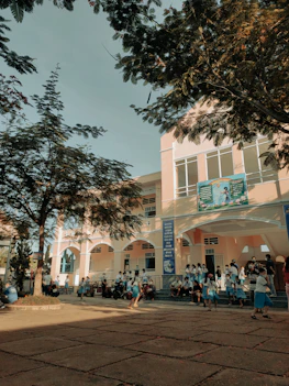 A group of children wearing school uniforms are gathered outside a large building with peach-colored walls and white windows. Some children are sitting on the steps while others are engaged in playful activities around the paved courtyard. Tall trees with dense foliage provide shade, casting intricate shadows on the ground.