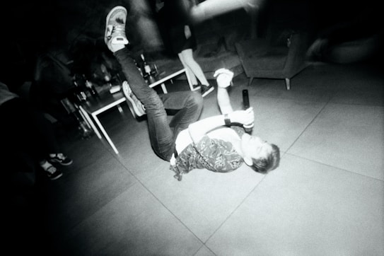 A person is lying on the floor holding a microphone, surrounded by a dynamic and energetic environment. There are tables with bottles and other people in the background, suggesting a party or social gathering. The scene is captured in monochrome, which adds a dramatic and gritty feel.