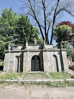 An old, weathered stone mausoleum with inscriptions and a cross on top, surrounded by lush green trees and sparse wildflowers growing at its base. The mausoleum has an arched entrance, and there are additional grave markers visible in the background.