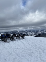 Several snowmobiles are parked on a snowy mountain landscape with a cloudy sky overhead. The distant view reveals snow-covered mountains under thick, gray clouds.
