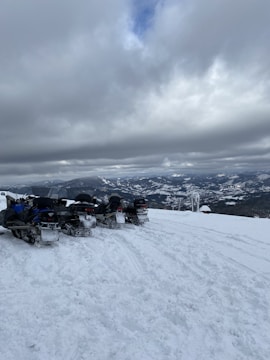 Several snowmobiles are parked on a snowy mountain landscape with a cloudy sky overhead. The distant view reveals snow-covered mountains under thick, gray clouds.