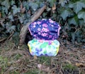 Two cloth diapers with colorful floral patterns are placed on a background of green ivy leaves and dried twigs. The top diaper has a navy blue background with pink cherry blossom designs, while the bottom diaper features a white background with multicolored flowers.