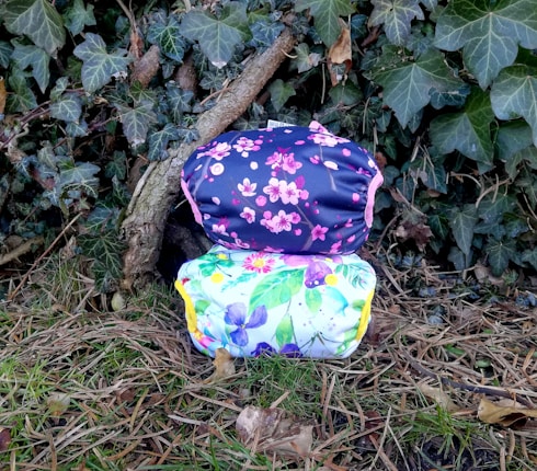 Two cloth diapers with colorful floral patterns are placed on a background of green ivy leaves and dried twigs. The top diaper has a navy blue background with pink cherry blossom designs, while the bottom diaper features a white background with multicolored flowers.