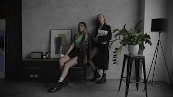 Two women pose in a minimalist room with a light gray textured wall. One is sitting on a black console table adorned with a framed abstract artwork and two candles. She wears a green crop top, black shorts, and a leather jacket. The other stands beside a large potted plant and holds a magazine, dressed in a long black coat. A modern black floor lamp with a slender frame stands next to the plant.