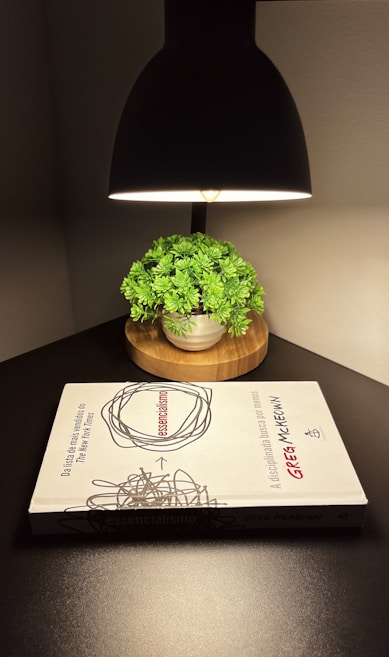An overhead lamp illuminates a small green plant in a pot placed on a wooden base. In front of the plant, there is a book titled 'Essencialismo' by Greg McKeown on a dark tabletop.
