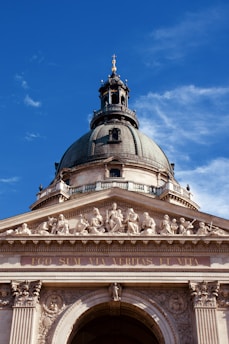 A grand architectural structure featuring a large dome with a cross at the top, intricate sculptures of figures along the frieze, and the Latin inscription 'EGO SUM VIA VERITAS ET VITA' on the front. The structure is made of stone with detailed carvings and is set against a clear blue sky.