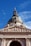 A grand architectural structure featuring a large dome with a cross at the top, intricate sculptures of figures along the frieze, and the Latin inscription 'EGO SUM VIA VERITAS ET VITA' on the front. The structure is made of stone with detailed carvings and is set against a clear blue sky.