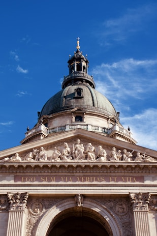 A grand architectural structure featuring a large dome with a cross at the top, intricate sculptures of figures along the frieze, and the Latin inscription 'EGO SUM VIA VERITAS ET VITA' on the front. The structure is made of stone with detailed carvings and is set against a clear blue sky.