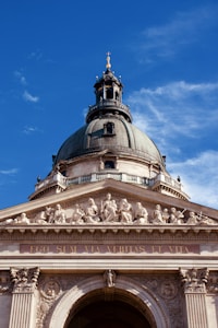 A grand architectural structure featuring a large dome with a cross at the top, intricate sculptures of figures along the frieze, and the Latin inscription 'EGO SUM VIA VERITAS ET VITA' on the front. The structure is made of stone with detailed carvings and is set against a clear blue sky.