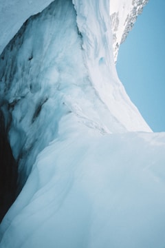 A close-up view of a glacier with smooth, flowing icy surfaces and crevasses. The ice appears blue and white, with sunlight reflecting off its textured curves. In the background, there is a clear blue sky contrasting with the ice.