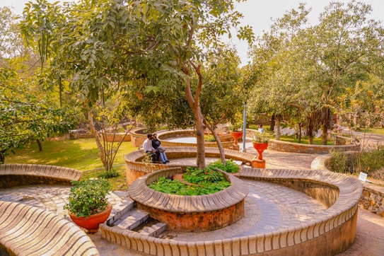 A verdant park with winding pathways and circular seating arrangements constructed from bricks and concrete. Trees and plants are abundant throughout the area, and there are a few potted plants placed strategically. Two people are sitting on one of the circular benches, possibly engaged in conversation. The atmosphere appears peaceful and inviting, with the sunlight casting a warm glow over the entire scene.