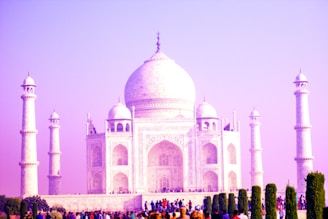 The iconic Taj Mahal is captured with its white marble facade glowing under a pink-hued sky. Four minarets stand gracefully at each corner, and intricate architectural details are visible on the main structure. A crowd of visitors in colorful clothing gathers at the base, surrounded by lush green gardens.