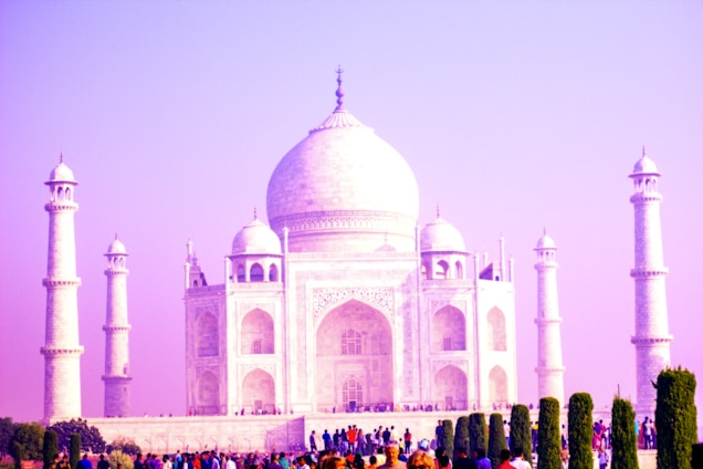 The iconic Taj Mahal is captured with its white marble facade glowing under a pink-hued sky. Four minarets stand gracefully at each corner, and intricate architectural details are visible on the main structure. A crowd of visitors in colorful clothing gathers at the base, surrounded by lush green gardens.