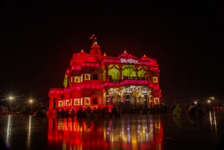 A richly illuminated temple stands majestically against a night sky, its intricate architecture highlighted by vibrant red and yellow lighting. The reflection of the lights casts dramatic patterns on the surrounding polished surface. A few silhouetted figures are visible seated near the structure, enhancing the serene and spiritual atmosphere.