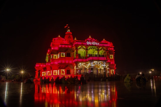 A richly illuminated temple stands majestically against a night sky, its intricate architecture highlighted by vibrant red and yellow lighting. The reflection of the lights casts dramatic patterns on the surrounding polished surface. A few silhouetted figures are visible seated near the structure, enhancing the serene and spiritual atmosphere.