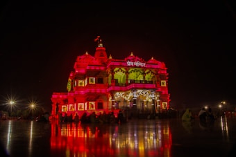 A richly illuminated temple stands majestically against a night sky, its intricate architecture highlighted by vibrant red and yellow lighting. The reflection of the lights casts dramatic patterns on the surrounding polished surface. A few silhouetted figures are visible seated near the structure, enhancing the serene and spiritual atmosphere.