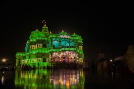A brightly illuminated temple adorned with green lights stands majestically at night. The intricate architecture features multiple tiers and spires, with details highlighted by the vibrant lighting. In front of the temple, a crowd of people is gathered, some sitting and some standing, casting soft reflections on the polished ground.