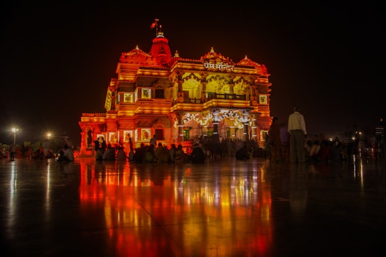 An ornate, illuminated temple glows in vibrant shades of orange and red against a dark night sky. Reflections on the glossy floor enhance the vibrant colors. People are gathered around the temple, adding a sense of community and reverence to the scene.