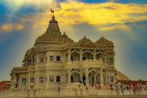 A magnificent temple with intricate architecture is set against a dynamic sky with dramatic cloud formations. The temple is adorned with detailed carvings and sculptures, showcasing exquisite craftsmanship. People can be seen walking around the temple, adding a sense of scale and activity.