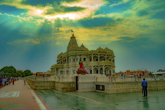 A grand temple with intricate carvings and multiple spires is bathed in sunlight streaming through the clouds. The scene includes people walking around the temple on the spacious pavement, enhancing the temple's majestic appearance.