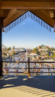 A vibrant outdoor scene with numerous people walking and socializing in an open plaza. The area is bordered by buildings with distinct architectural styles. The plaza's surface reflects the sunlight, creating an inviting atmosphere. In the background, lush trees and rooftops are visible under a clear blue sky.