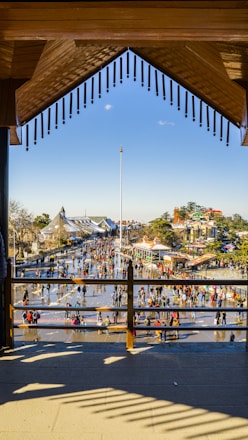 A vibrant outdoor scene with numerous people walking and socializing in an open plaza. The area is bordered by buildings with distinct architectural styles. The plaza's surface reflects the sunlight, creating an inviting atmosphere. In the background, lush trees and rooftops are visible under a clear blue sky.