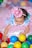 A baby dressed in a colorful, whimsical outfit featuring blue and pink colors and adorned with pom-poms plays among a variety of multicolored plastic balls. The child wears a small, sparkly pink hat that matches the playful theme of the outfit. The background is filled with more soft and pastel-colored balls, adding to the festive atmosphere.