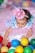 A baby dressed in a colorful, whimsical outfit featuring blue and pink colors and adorned with pom-poms plays among a variety of multicolored plastic balls. The child wears a small, sparkly pink hat that matches the playful theme of the outfit. The background is filled with more soft and pastel-colored balls, adding to the festive atmosphere.