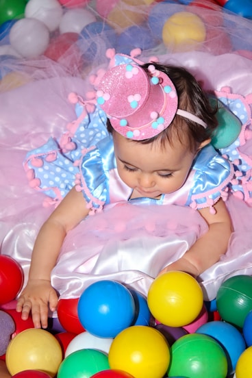 A baby dressed in a colorful, whimsical outfit featuring blue and pink colors and adorned with pom-poms plays among a variety of multicolored plastic balls. The child wears a small, sparkly pink hat that matches the playful theme of the outfit. The background is filled with more soft and pastel-colored balls, adding to the festive atmosphere.
