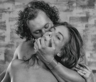 A black and white photograph featuring two individuals in a close, intimate embrace. The man gently holds the woman's face with one hand while kissing her. Both have relaxed expressions, and the woman has long hair that cascades over her shoulders. The background consists of an abstract, textured pattern.